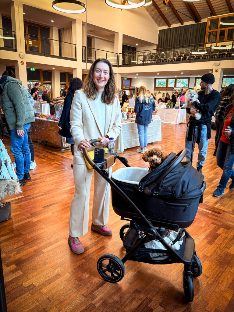 A woman at an indoor market pushing a small brown poodle in a black dog stroller, surrounded by shoppers and handmade stalls.
