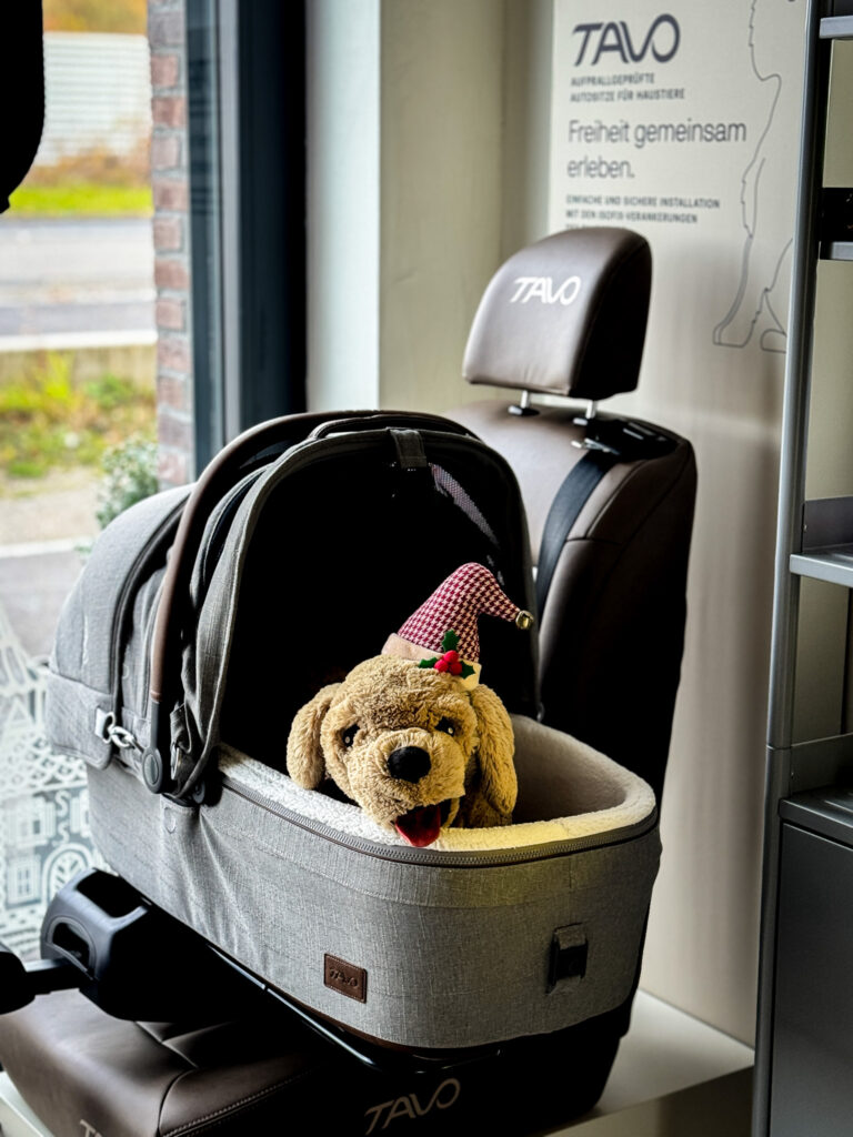 A grey dog car seat on display with a stuffed dog toy inside wearing a festive holiday hat.