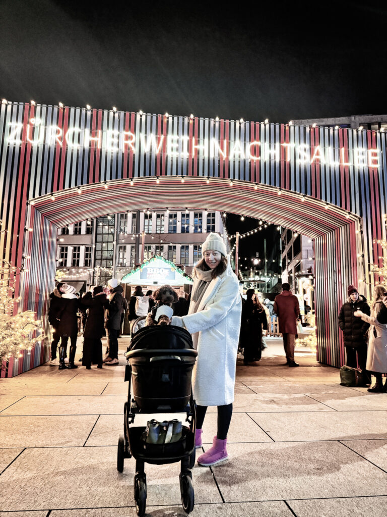 A woman smiling beside a stroller with her small dog at the illuminated entrance of the Weihnachtsallee Christmas Market in Zurich.