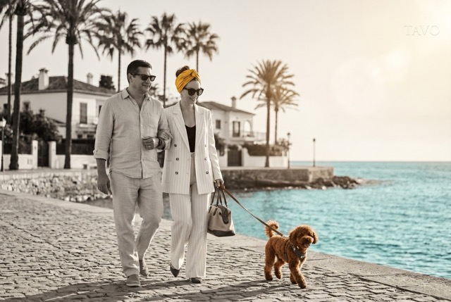 Stylish couple walking a red toy poodle along a palm-lined seaside promenade on holiday