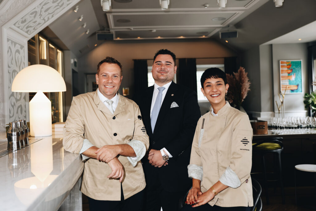 Three hospitality professionals, two in beige bar jackets and one in a dark suit, standing at a white marble bar