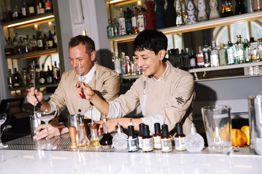 Two smiling bartenders in beige jackets stirring cocktails side by side at a white marble bar, with bottles on lit shelves behind them